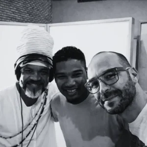 Fotografia em preto e branco de Carlinhos Brown com turbante branco, Iuri Passos e Wagner Barbosa sorrindo juntos em estúdio.