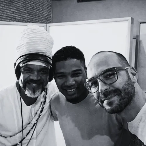 Fotografia em preto e branco de Carlinhos Brown com turbante branco, Iuri Passos e Wagner Barbosa sorrindo juntos em estúdio.