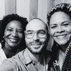 Fotografia em preto e branco em plano fechado de Margareth Menezes, Wagner Barbosa usando óculos e Márcia Short sorrindo juntos.