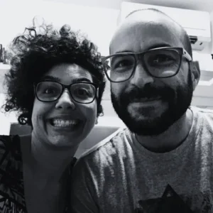 Fotografia em preto e branco de Vanessa Moreno sorrindo com cabelo cacheado e óculos, ao lado de Wagner Barbosa, também usando óculos.