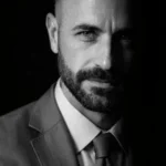 Foto de perfil em formato circular do preparador vocal Wagner Barbosa, vestindo terno e gravata, com iluminação dramática de estúdio em preto e branco.