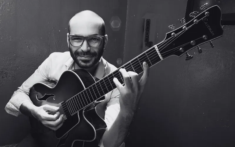 O preparador vocal e músico Wagner Barbosa sorrindo enquanto toca violão de guitarra semiacústica em foto em preto e branco.