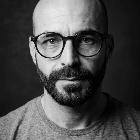Fotografia em preto e branco em close-up de Wagner Barbosa, preparador vocal e cantor, usando óculos de armação redonda e barba.